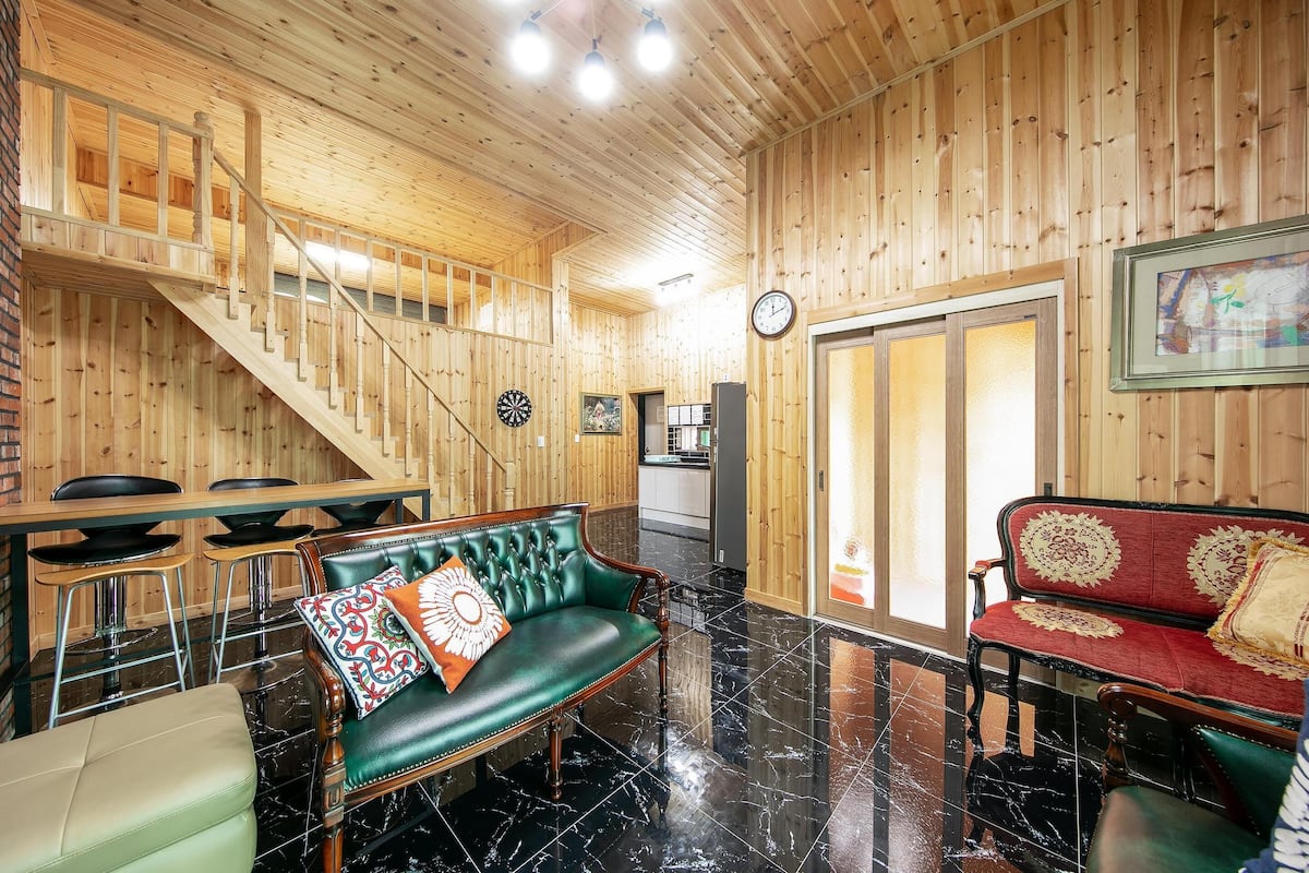 A wooden interior with a staircase, a dining area, a green sofa, and a red sofa.