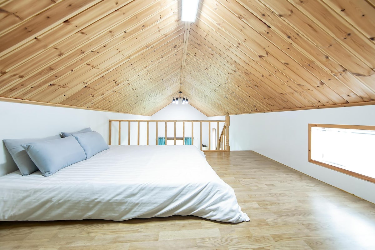 A bedroom with a sloped wooden ceiling, a bed with white bedding, and a large window on the right.