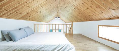 Quarto com teto de madeira inclinado, cama com roupa de cama branca e uma janela grande à direita.