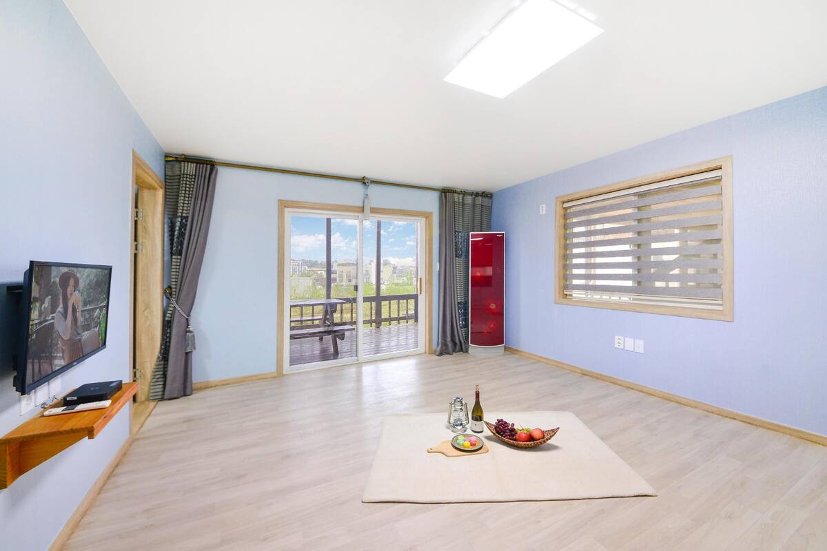 A spacious room with a large sliding glass door leading to an outdoor deck, a flat-screen TV mounted on the wall, and a small wooden shelf with a bottle and a bowl of fruit.