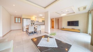 A spacious living area with a dining table, chairs, a coffee table with a decorative item, a TV on the wall, and a kitchen in the background. - Gapyeong Club Fish Villas Pension (Gapyeong)