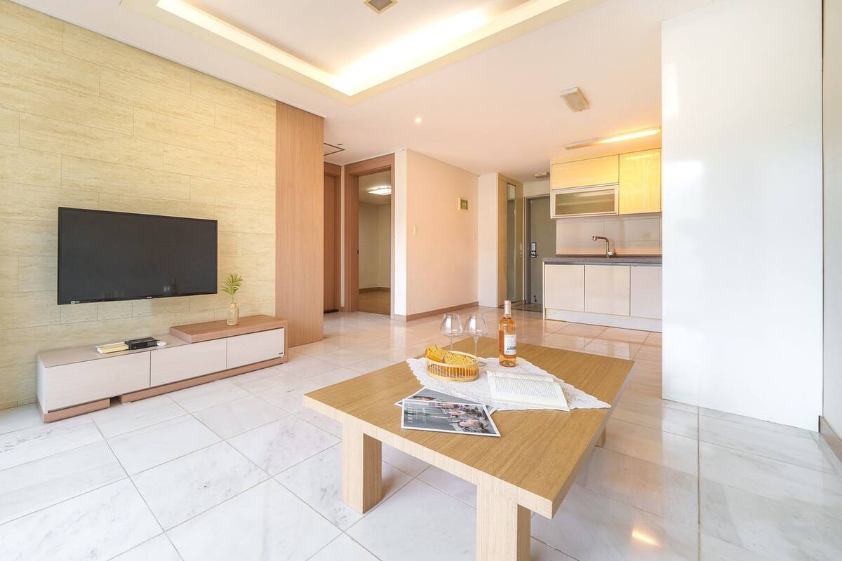 Una sala de estar moderna con una televisión de pantalla plana, una mesa de centro de madera y una cocina al fondo.