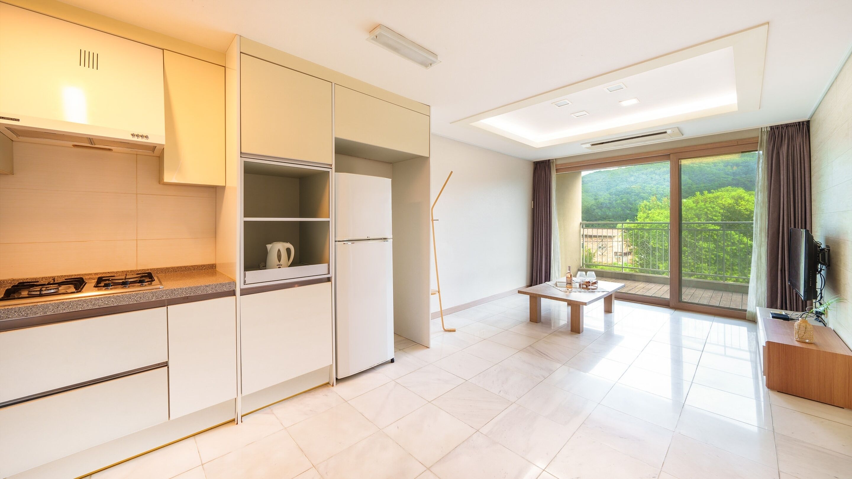 A modern kitchen with a gas stove, a kettle, and a refrigerator. There is a dining area with a table and chairs, and a balcony with a view of greenery.