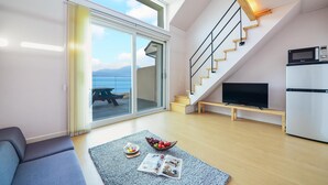 Un salon moderne avec un canapé, une table basse sur laquelle se trouve un magazine, un panier de fruits et un téléviseur posé sur un meuble en bois. Un escalier avec une rampe mène à un balcon d’où l’on a une vue sur la mer.