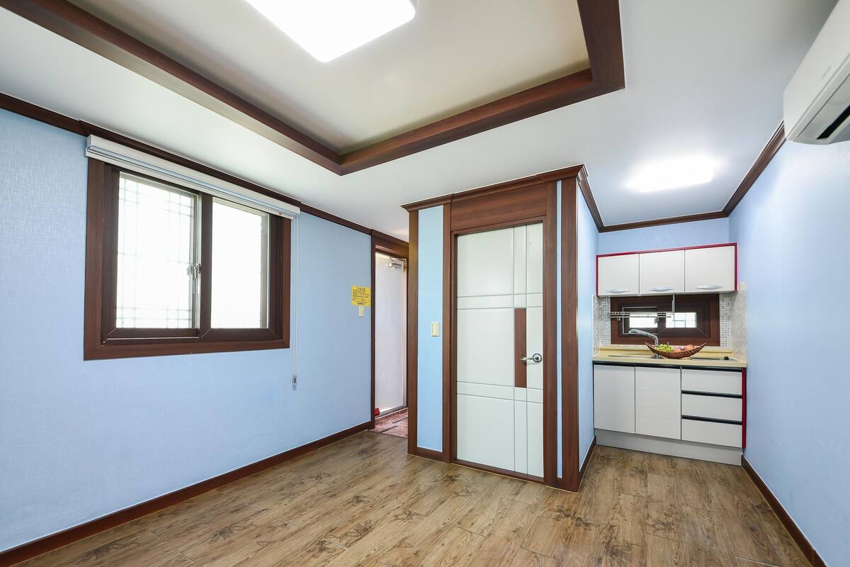A room with wooden flooring, a kitchen area with a sink and countertop, and a door leading to another room.