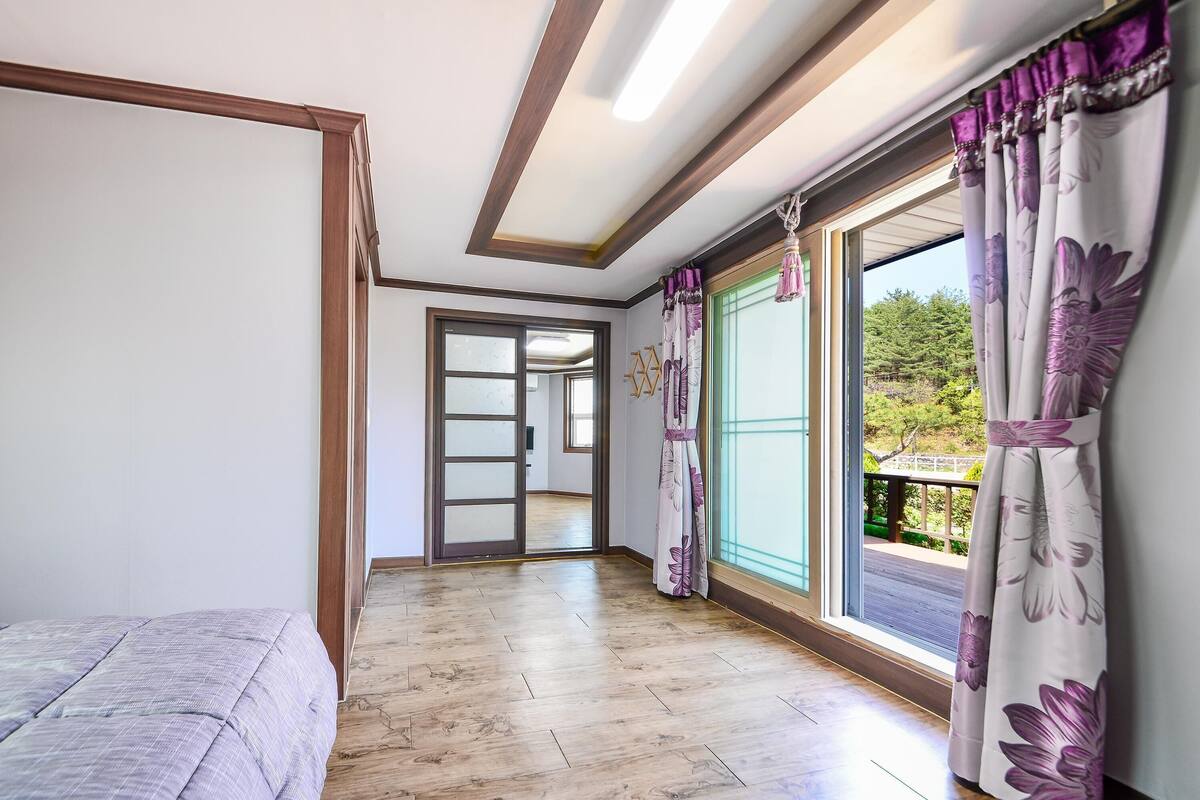 A room with a bed, a wooden floor, a large window with curtains, and a door leading to another room.