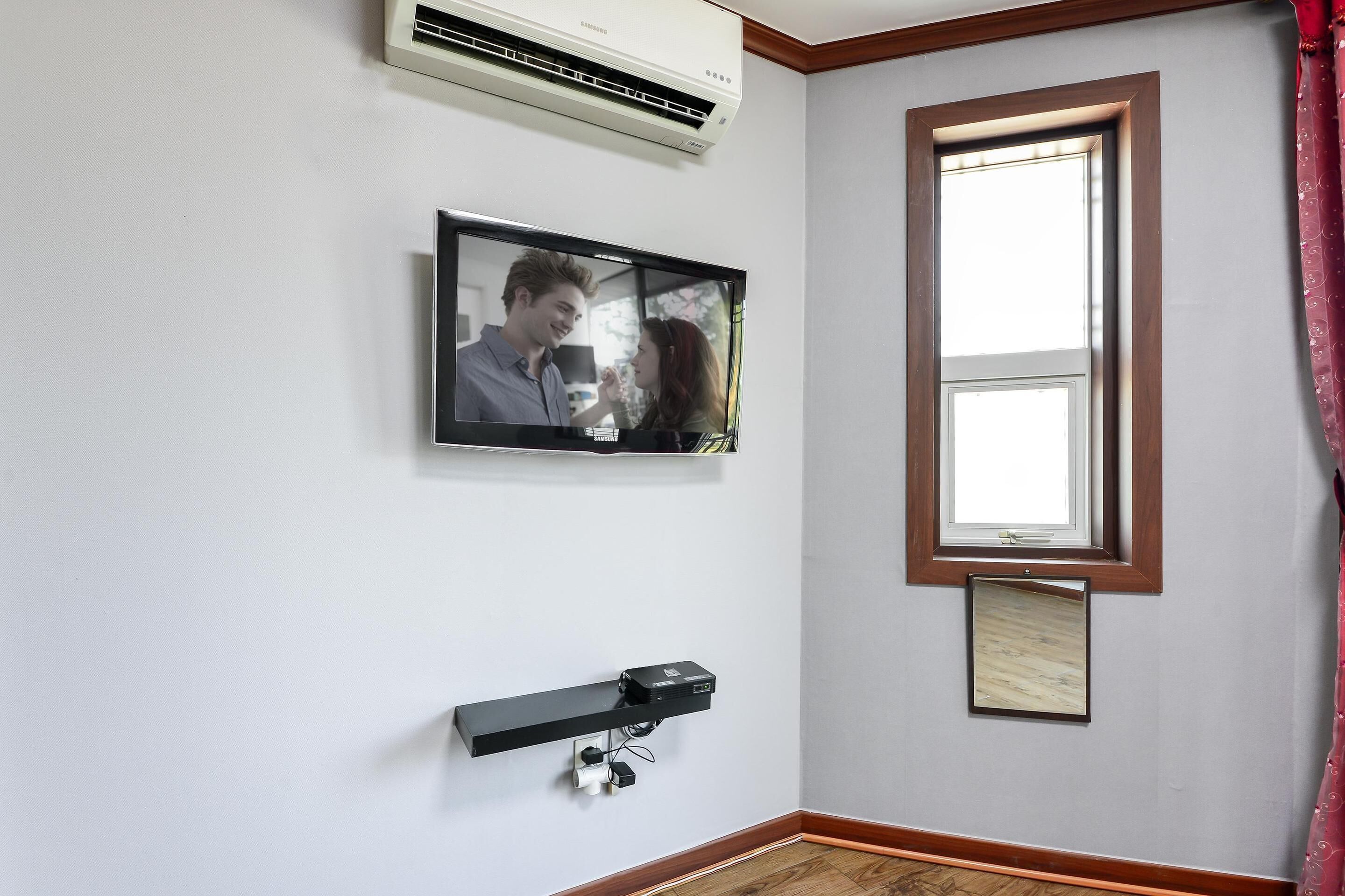 A room with a mounted TV displaying a couple, a window with a wooden frame, and a wall-mounted projector.