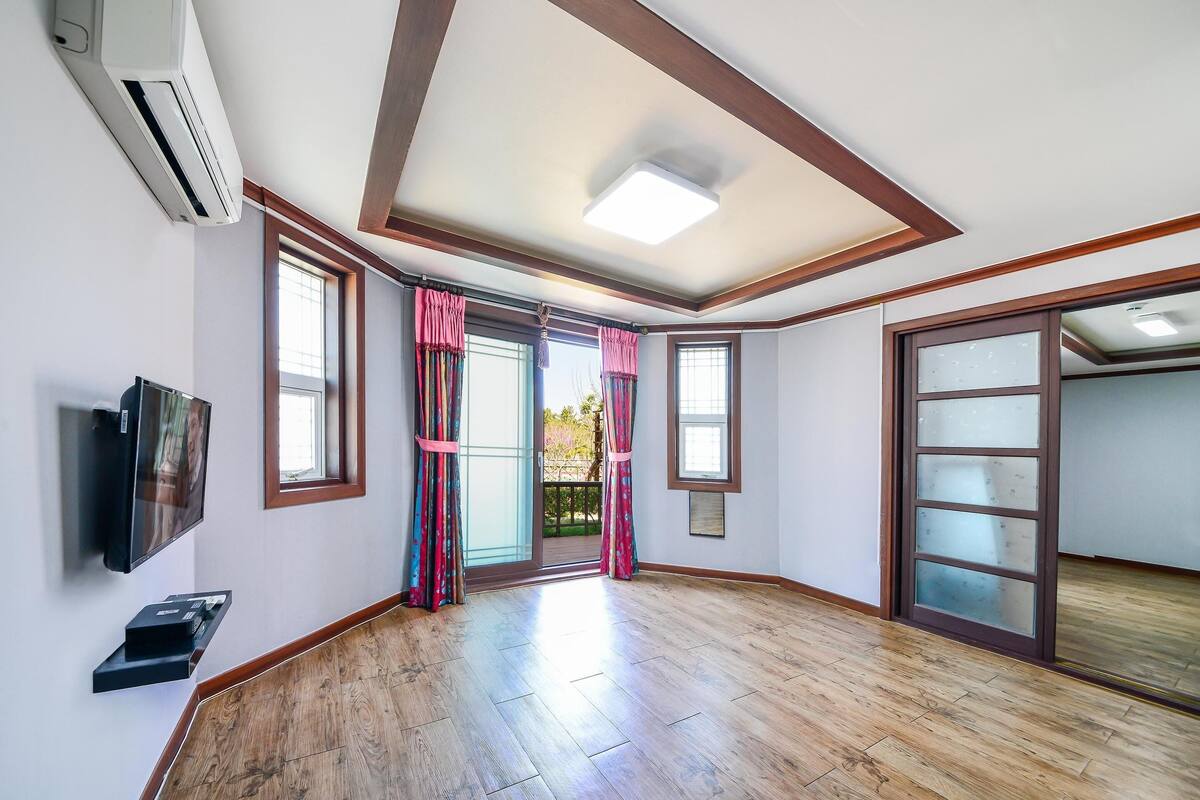 A spacious room with wooden flooring, a mounted TV, and a sliding glass door leading to a balcony.