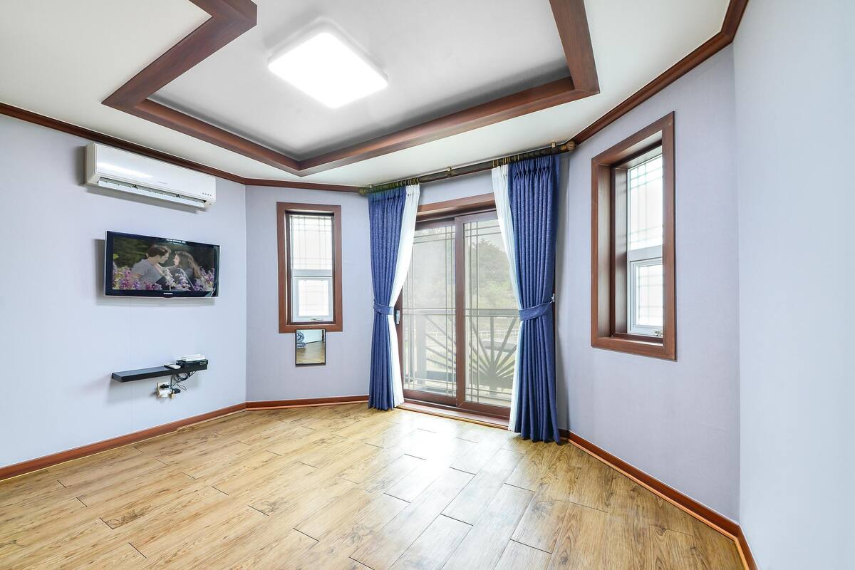 A room with wooden flooring, a ceiling fan, a mounted TV, and large windows with blue curtains.