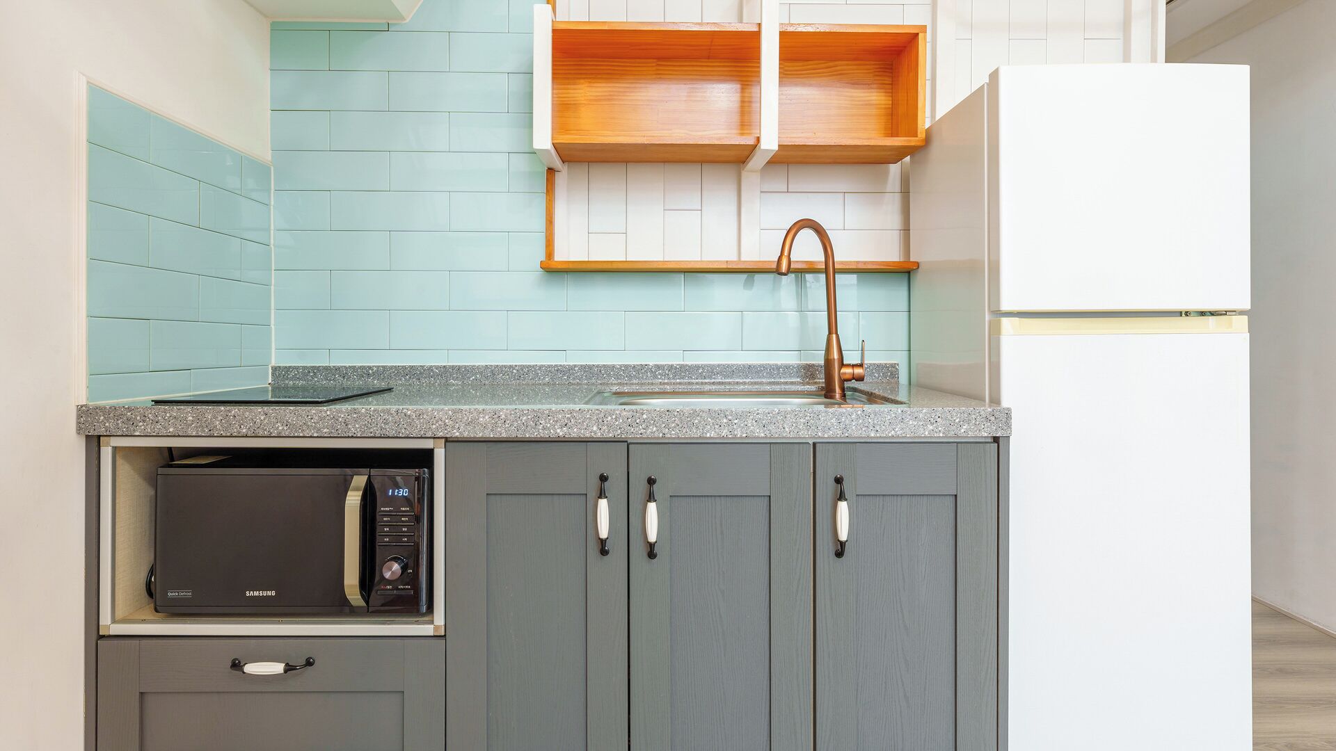 A kitchen with a built-in microwave, grey cabinets, a stainless steel sink, and a wooden shelf above the countertop.