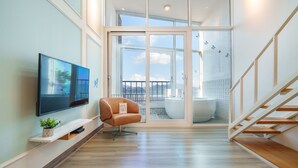 A modern living room with a flat-screen TV, a brown leather chair, a small potted plant, and a large sliding glass door leading to a balcony with a bathtub. - Yeosu Jplus Pension (Yeosu)