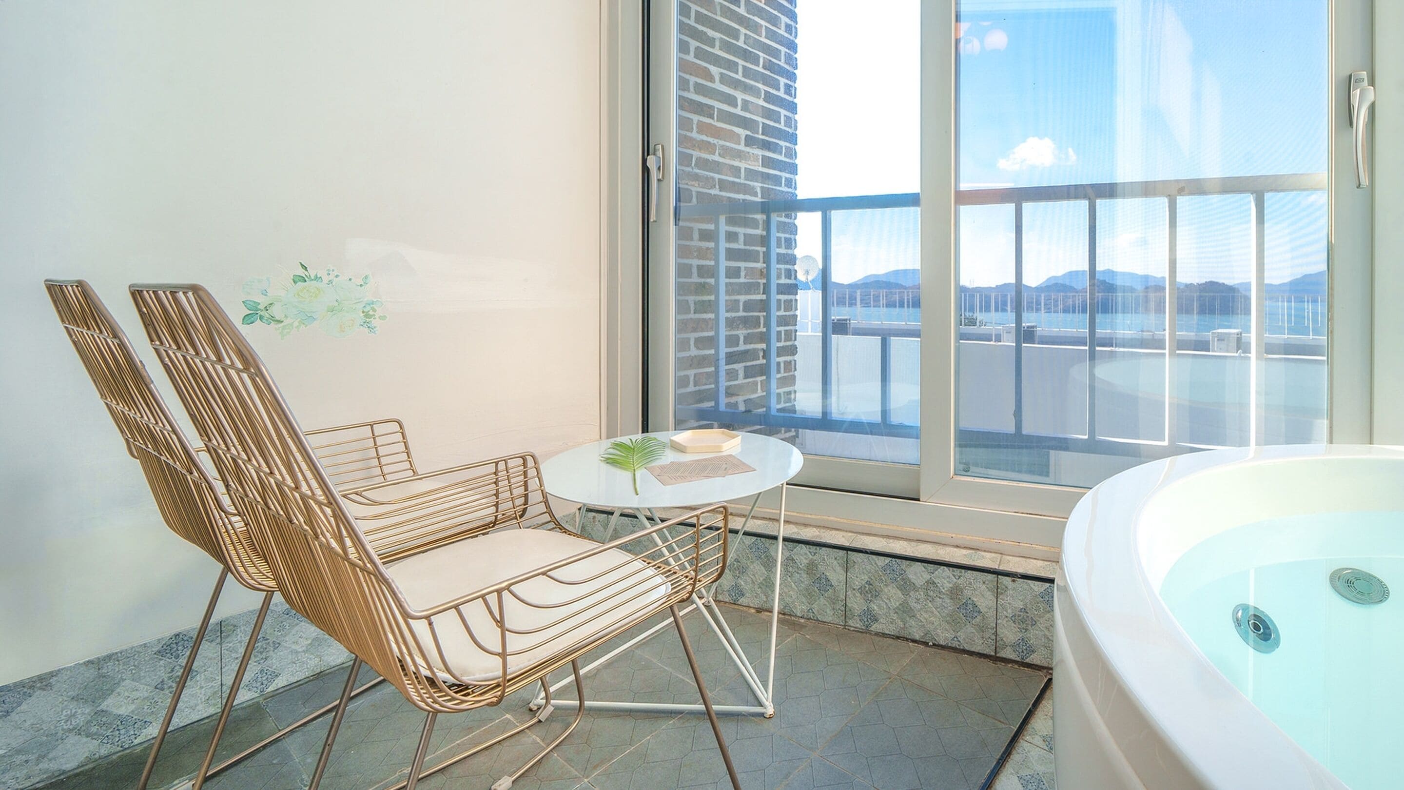 A bathroom with a bathtub, a wicker chair, a small table, and a large window with a view of a body of water and mountains.