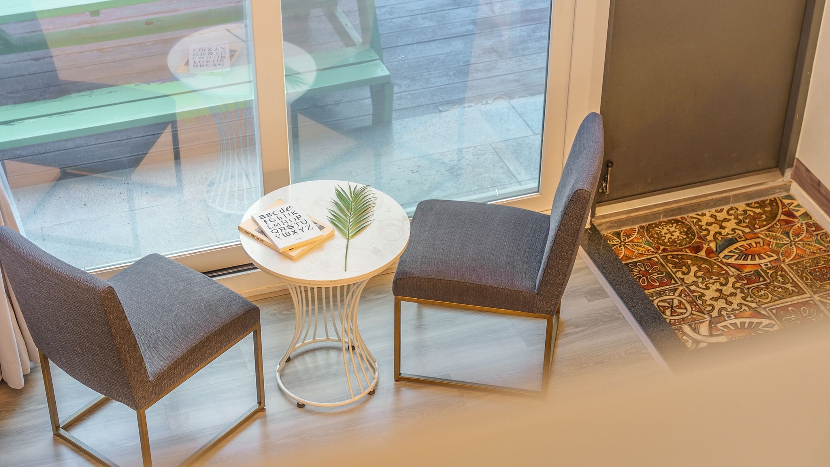 A room with a glass door, a small round table with a book and a leaf, two chairs, and a patterned rug.