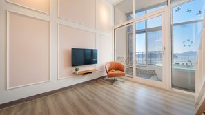 A modern living room with a flat-screen TV mounted on the wall, a wooden floor, and a large sliding glass door leading to a balcony with a view. - Yeosu Jplus Pension (Yeosu)