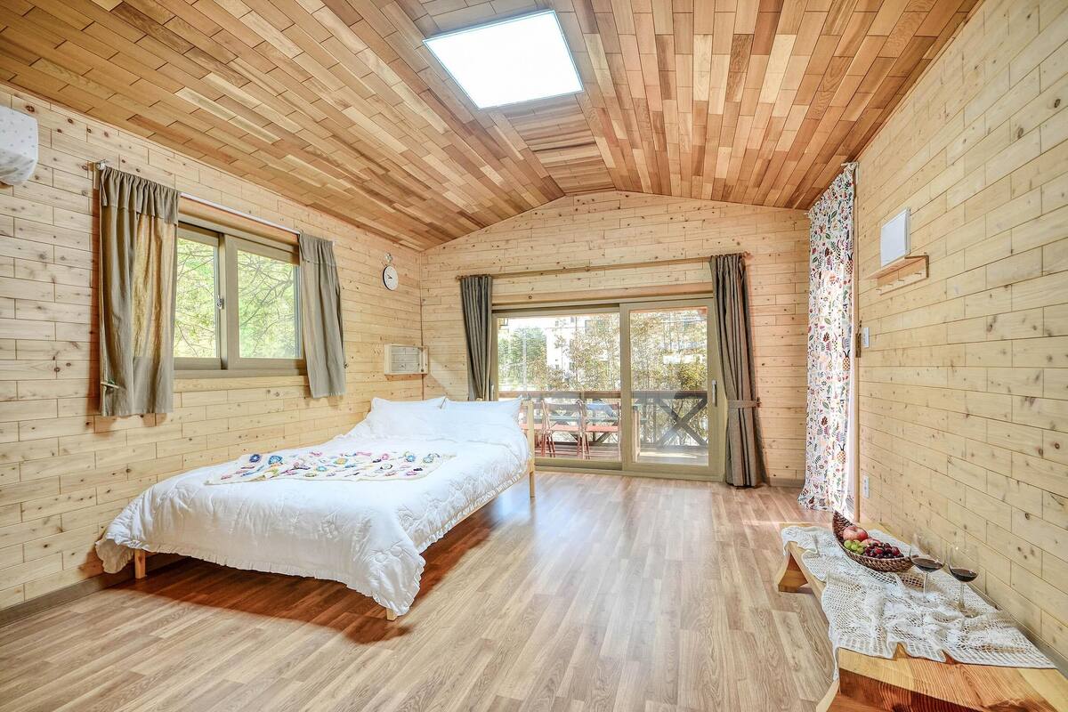 A wooden cabin bedroom with a bed, a table with fruit and wine glasses, and large windows.