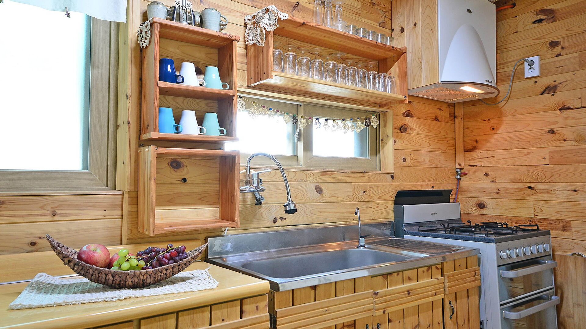 A wooden cabin-style kitchen with a sink, stove, and shelves with mugs and glasses.