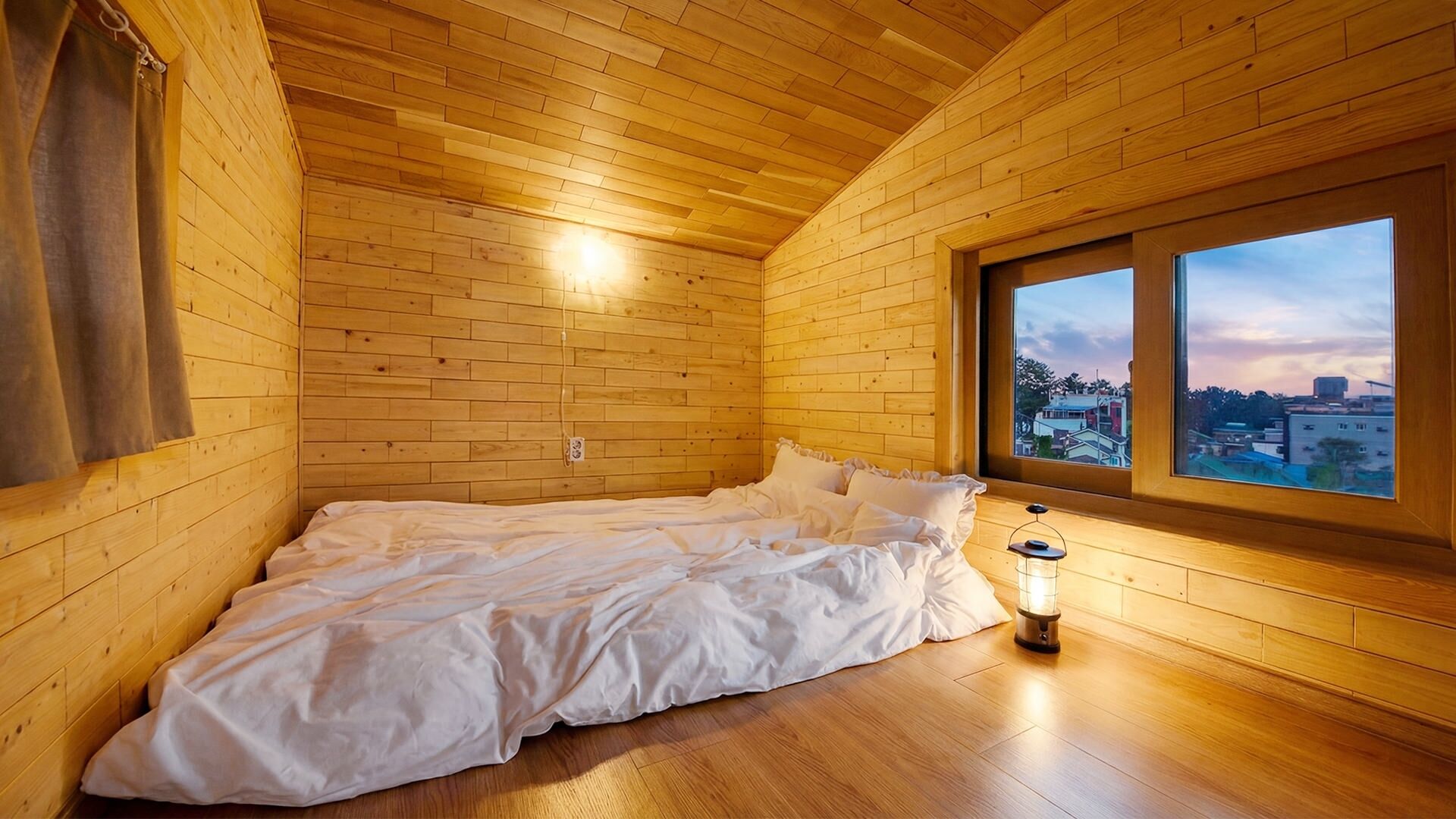 A single bed in a wooden room with a window overlooking a cityscape.