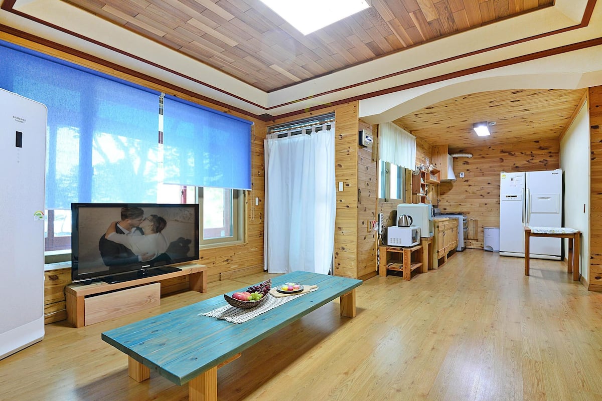 A cozy living area with a wooden ceiling, a television on a wooden stand, a dining table with a placemat, and a kitchen area with a refrigerator and microwave.