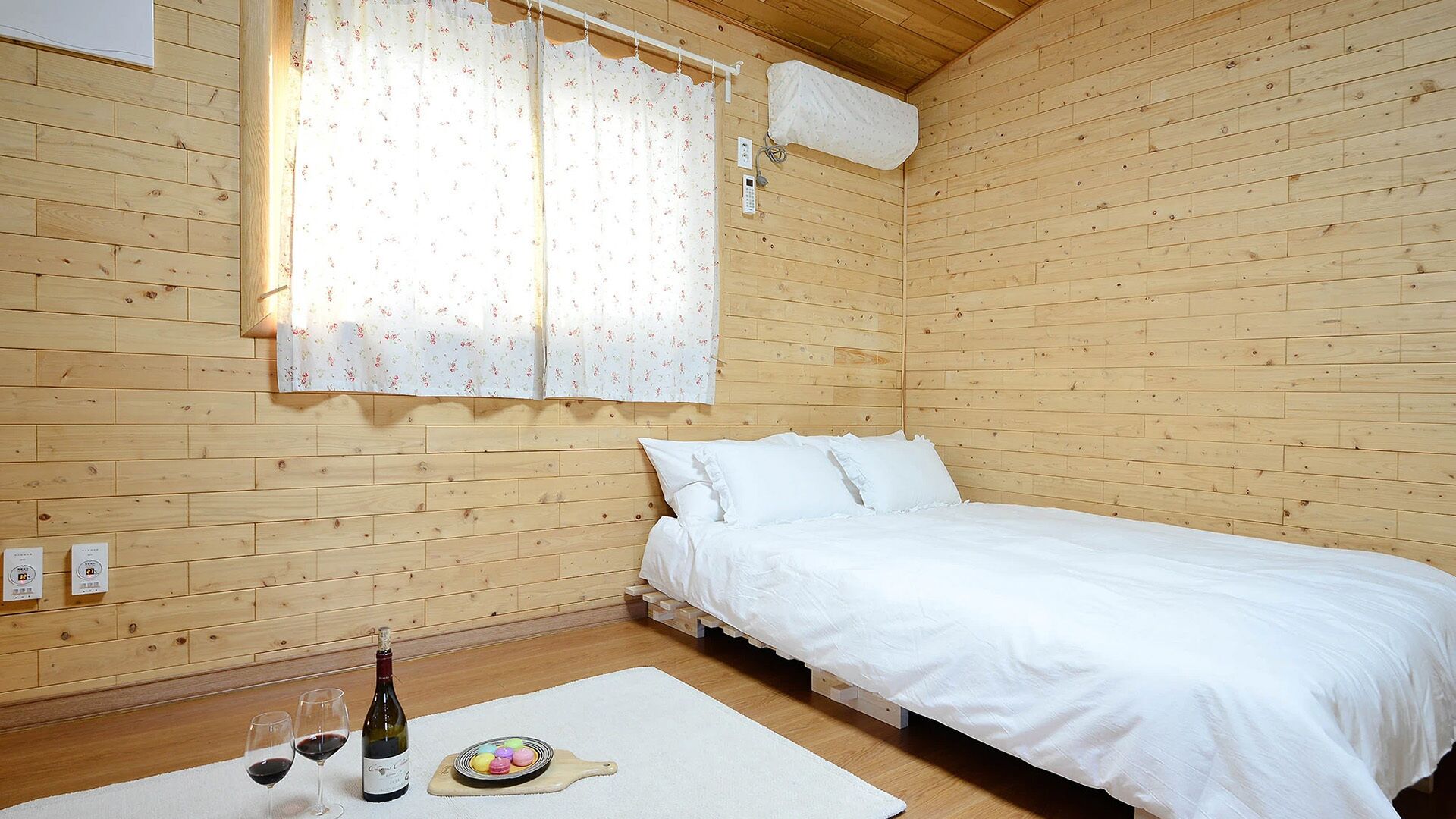Habitación de cabaña de madera con cama, ventana con cortinas, termostato y una botella de vino en el suelo.