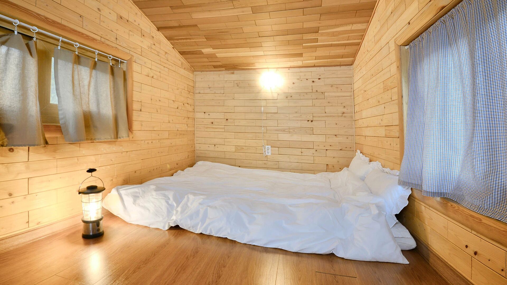 Habitación con paredes y techo de madera, cama con ropa blanca, lámpara colgante y ventana con cortinas.