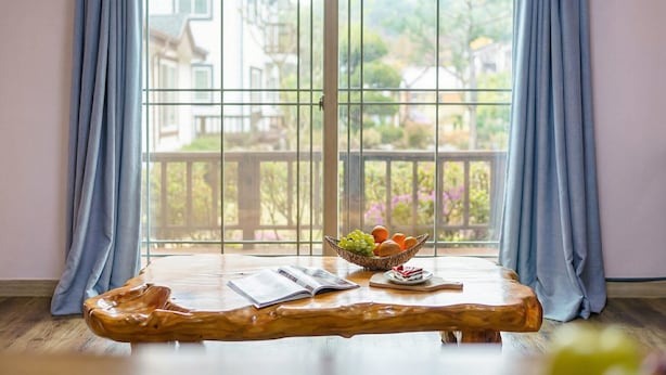 Una mesa de madera con un cuenco de fruta y un plato con una porción de pastel, ubicada frente a un amplio ventanal con vista a un balcón y edificios.