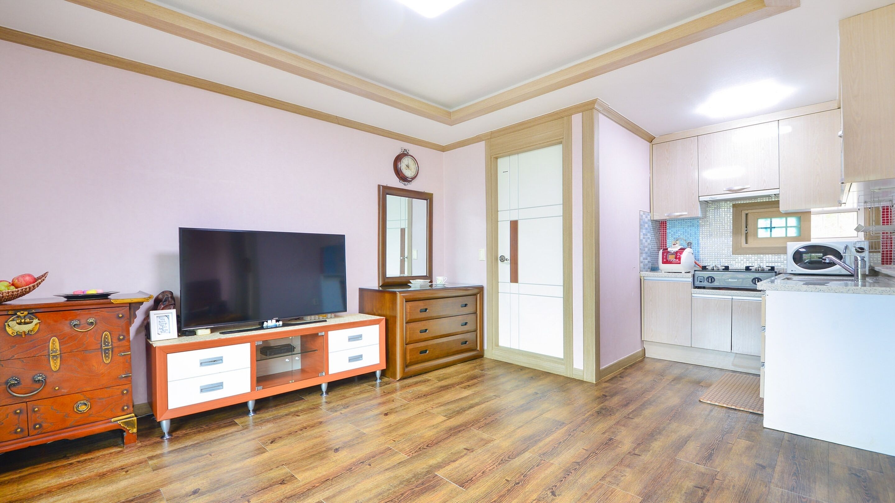 A compact living space with a TV, wooden dresser, and a kitchenette with a microwave and toaster.