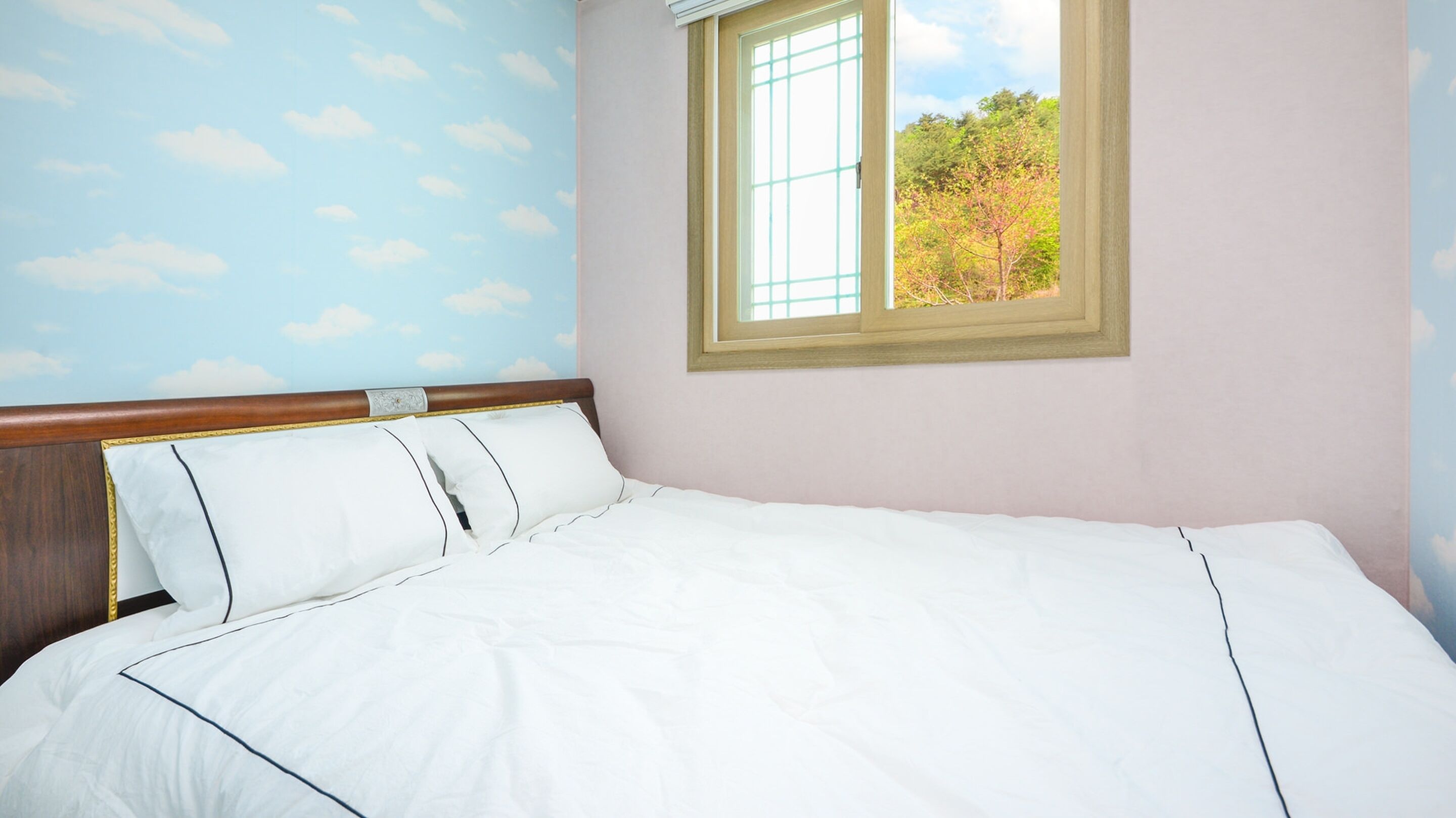 Una cama bien hecha con ropa blanca y un cabecero de madera, una ventana con vistas a árboles y una pared con un diseño de nubes.