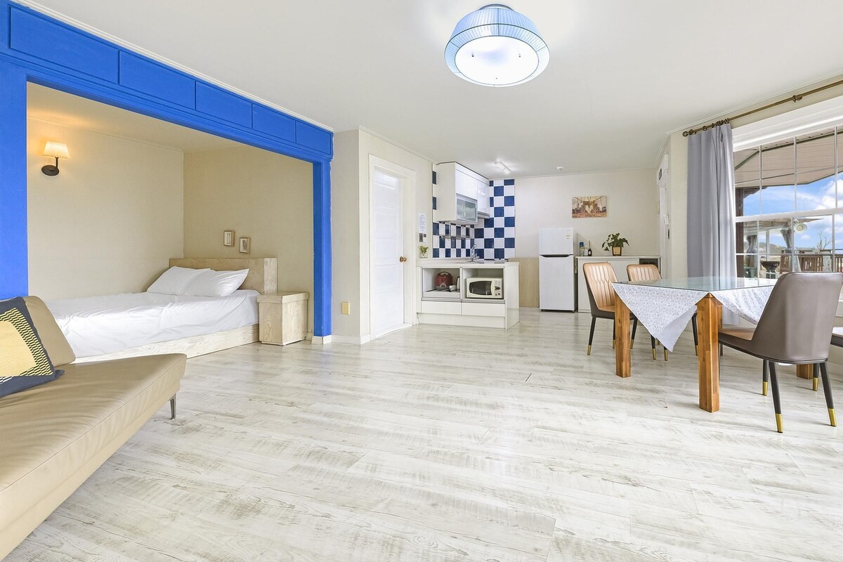 Une chambre d’hôtel moderne comprenant un lit, une petite kitchenette, un coin repas avec une table et des chaises, et une grande fenêtre avec des rideaux.