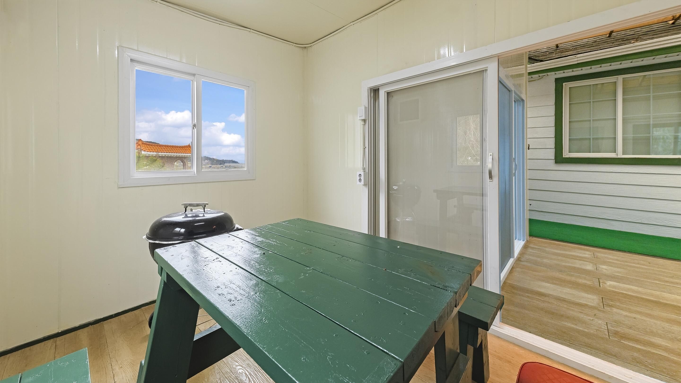 A small room with a green table and a barbecue grill. There is a window with a view of a building and a door leading to another room.