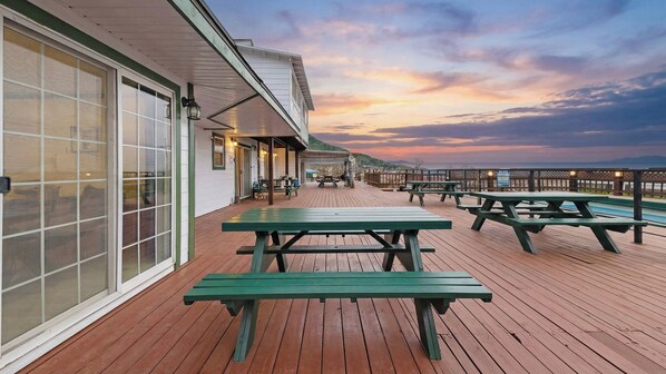 Una terraza de madera con mesas de picnic, un espacio de estar cubierto y vistas al atardecer sobre el agua.