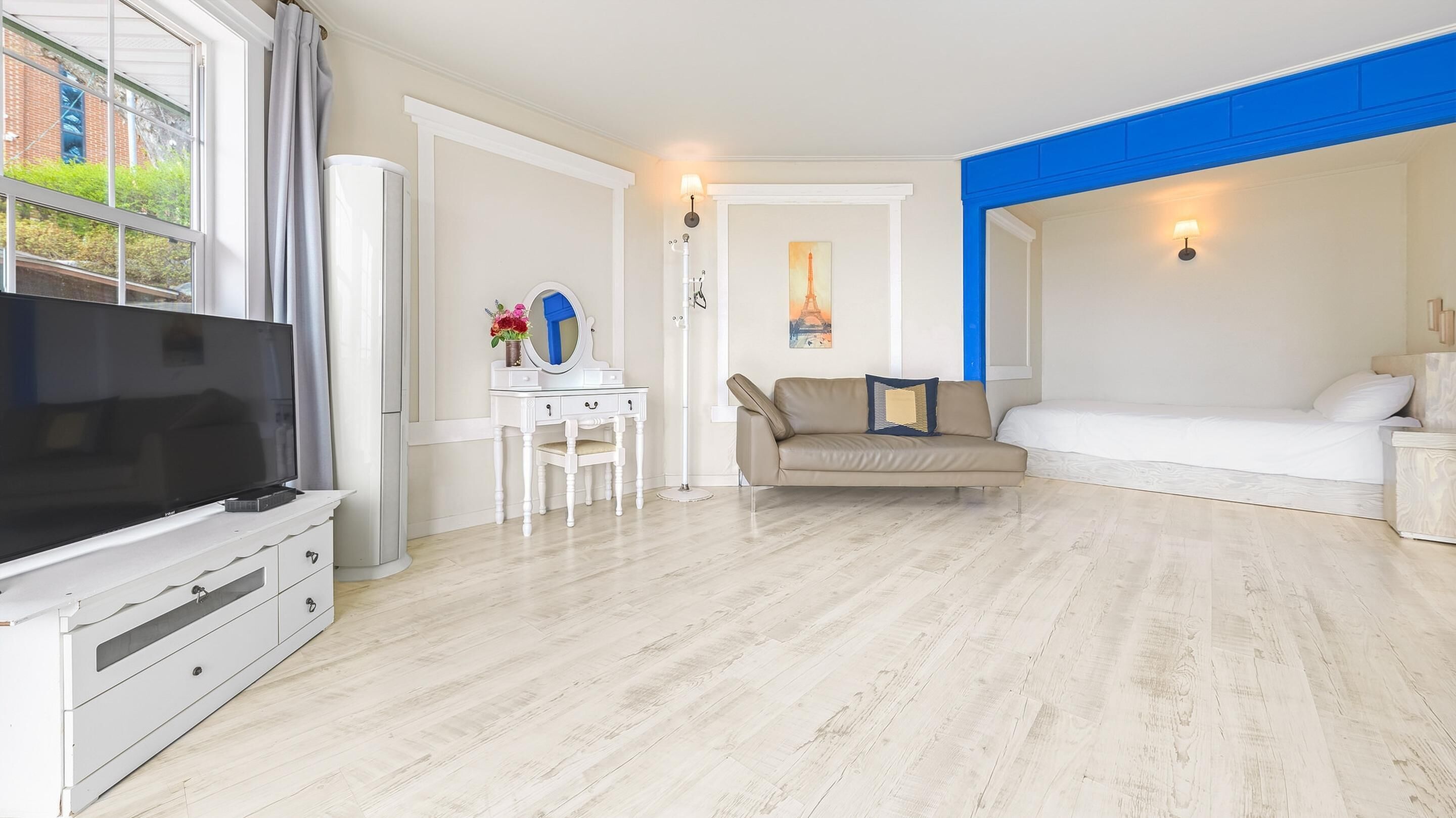 Uma sala de estar moderna com uma TV de tela plana, uma escrivaninha branca com uma cadeira, um sofá bege e uma cama branca.