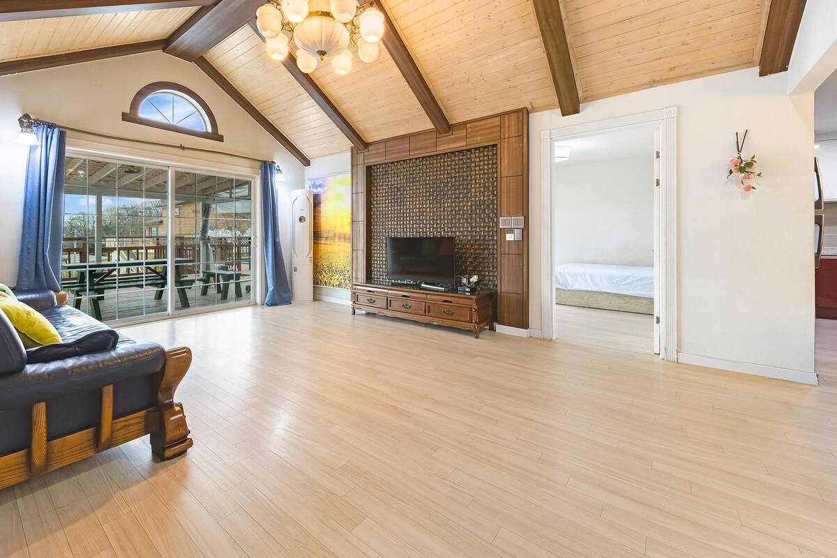 A spacious living room with a wooden ceiling, a large window, a sofa, a TV, and a wooden cabinet.