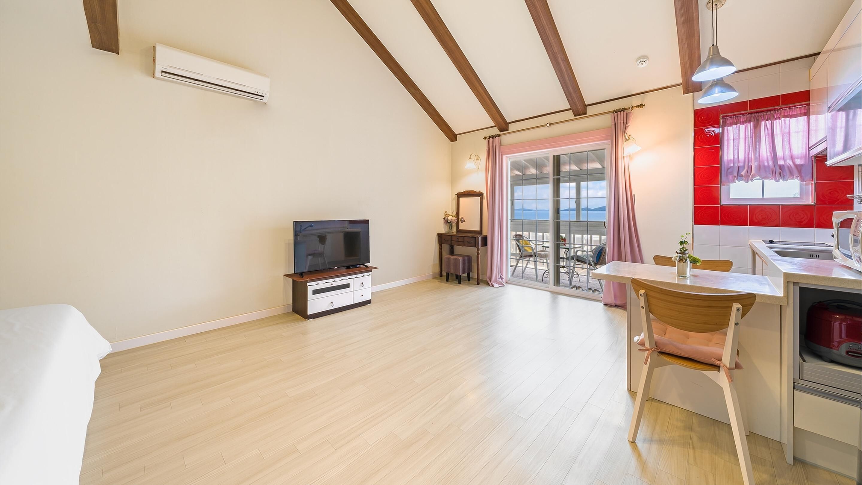 Una habitación amplia con piso de madera, un televisor sobre un mueble de madera, una zona de comedor con mesa y sillas, y un área de cocina con lavaplatos y estufa.