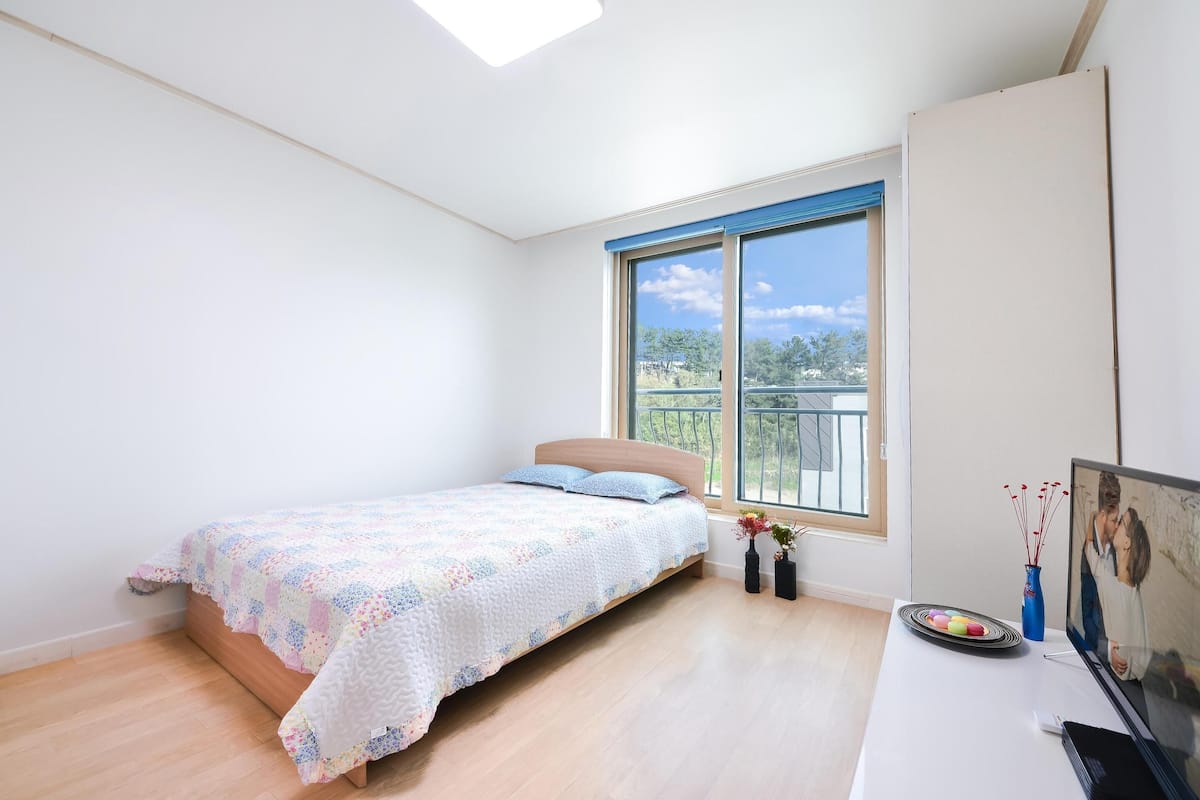 A bedroom with a bed, a desk, a TV, and a window with a view of trees.