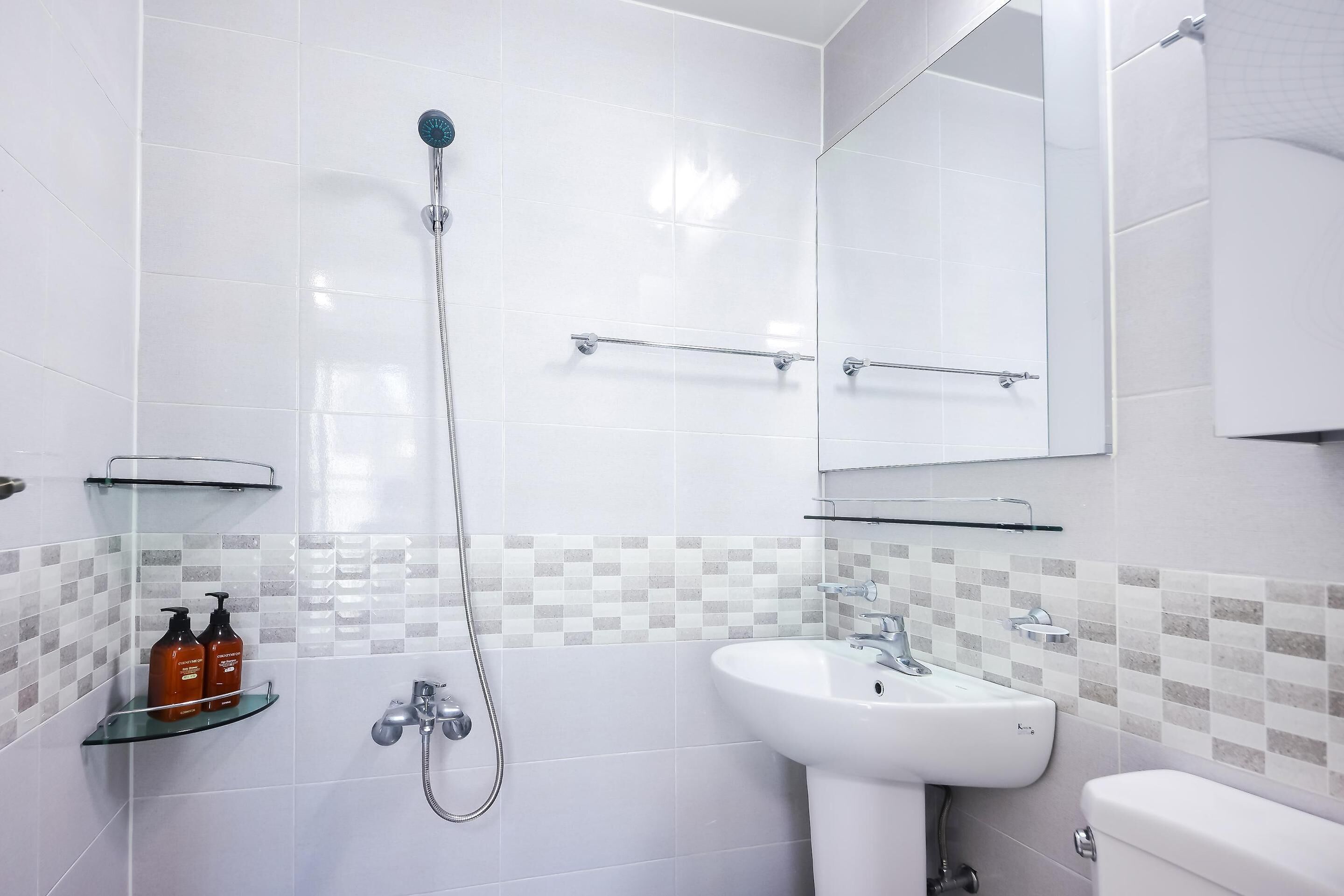 Et moderne badeværelse med en glasbrusekabine, en hvid vask og et toilet. To shampoo flasker står på en glashylde.