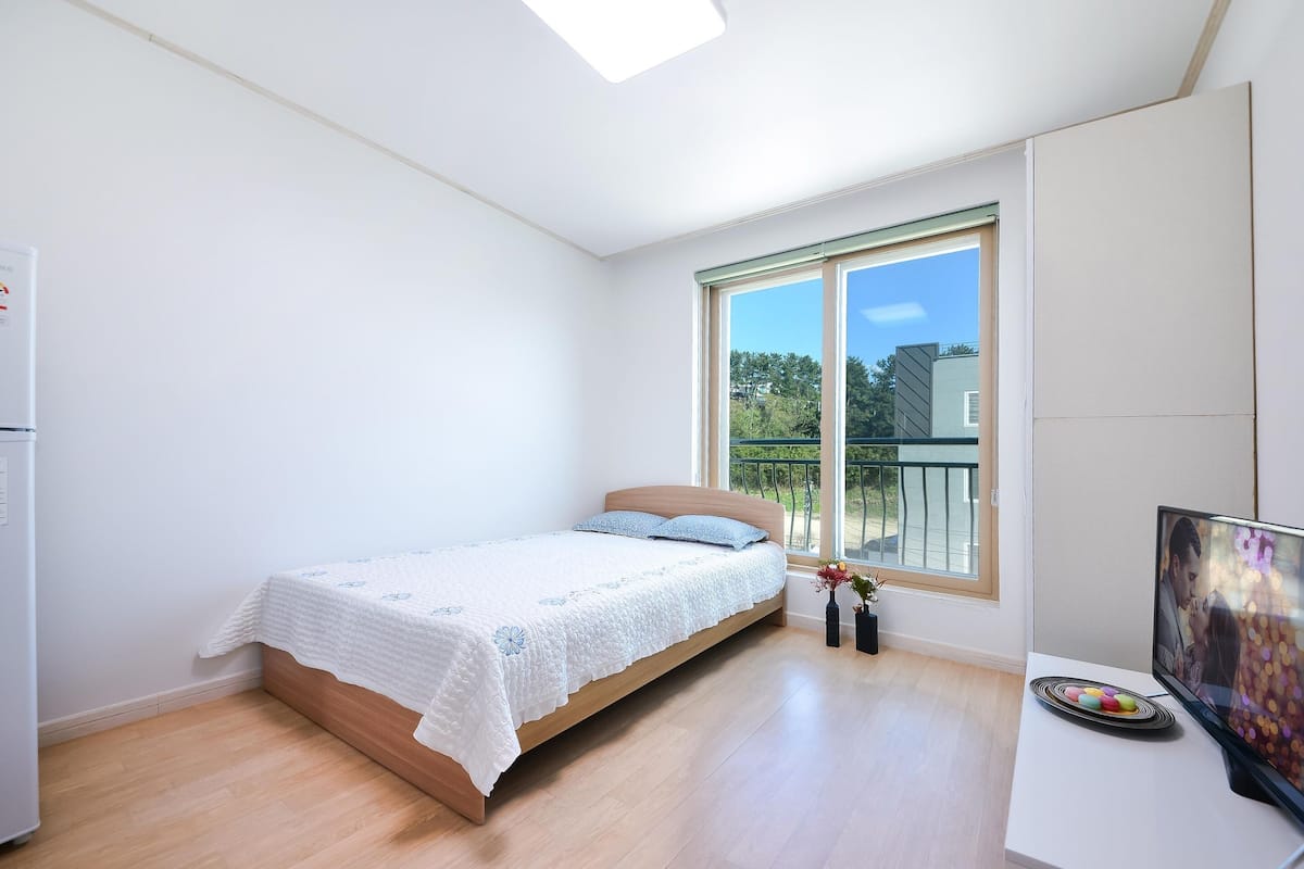 A bedroom with a bed, a TV, and a window with a view of greenery.