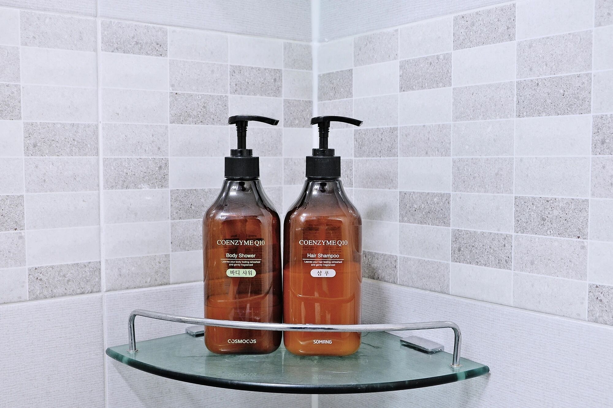 Two bottles of body shower and hair shampoo on a glass shelf in a bathroom with tiled walls.