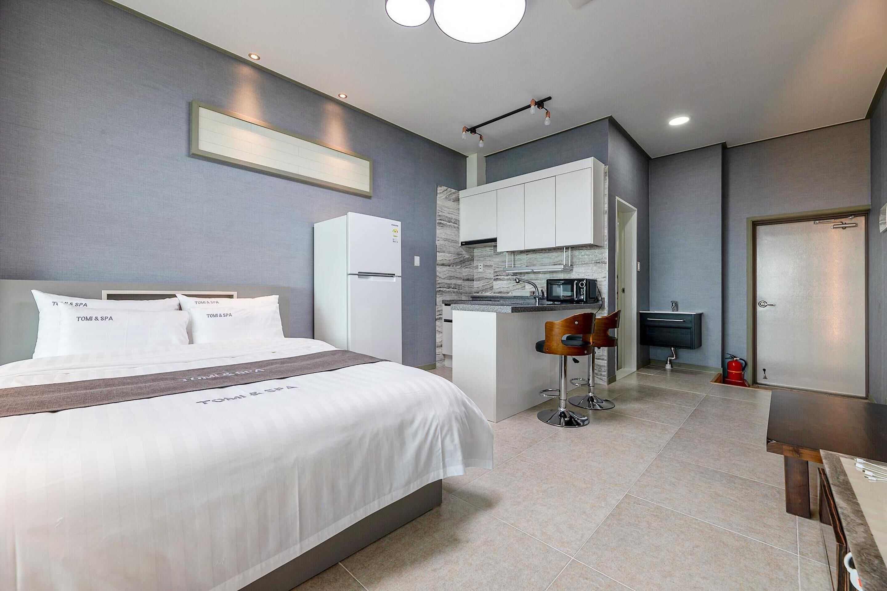 A modern hotel room with a large bed, a kitchenette, a small bar area, and a separate bathroom.