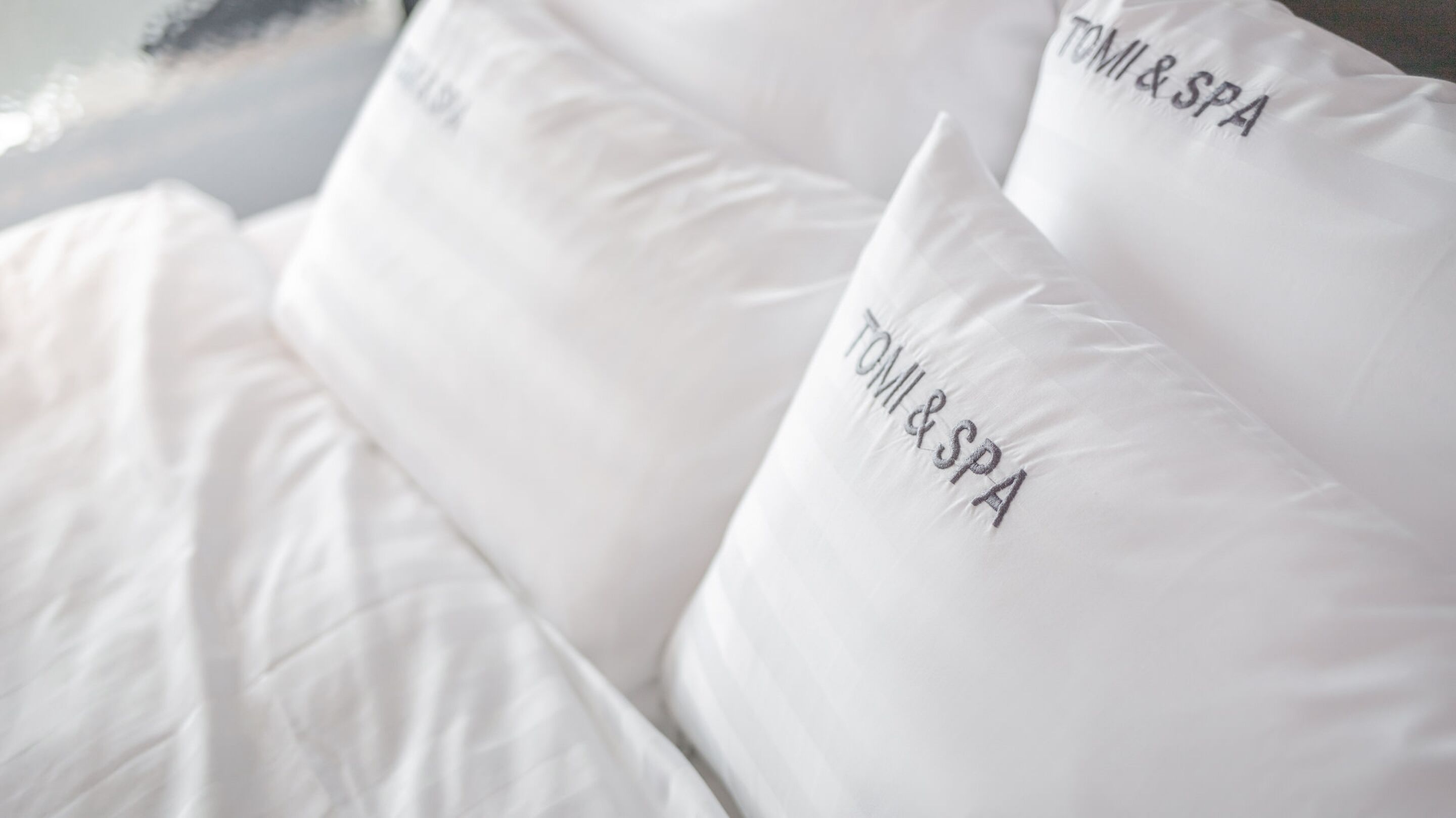A bed with white bedding and pillows that have "TOMI & SPA" embroidered on them.