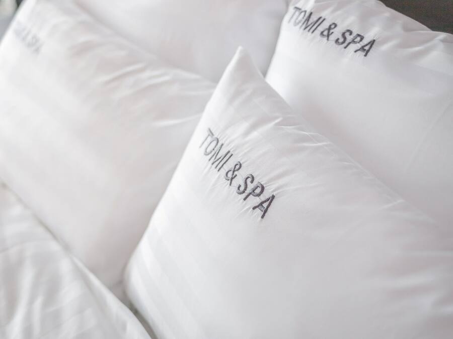 A bed with white bedding and pillows that have "TOMI & SPA" embroidered on them.