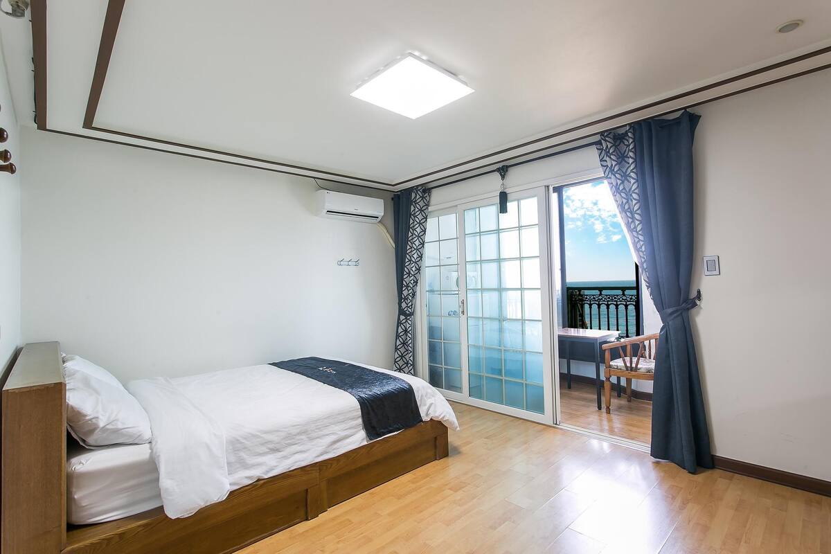 Une chambre avec un lit, un appareil de climatisation, un plancher en bois et une vue sur un espace extérieur aménagé avec une table et des chaises, accessible par une porte vitrée.