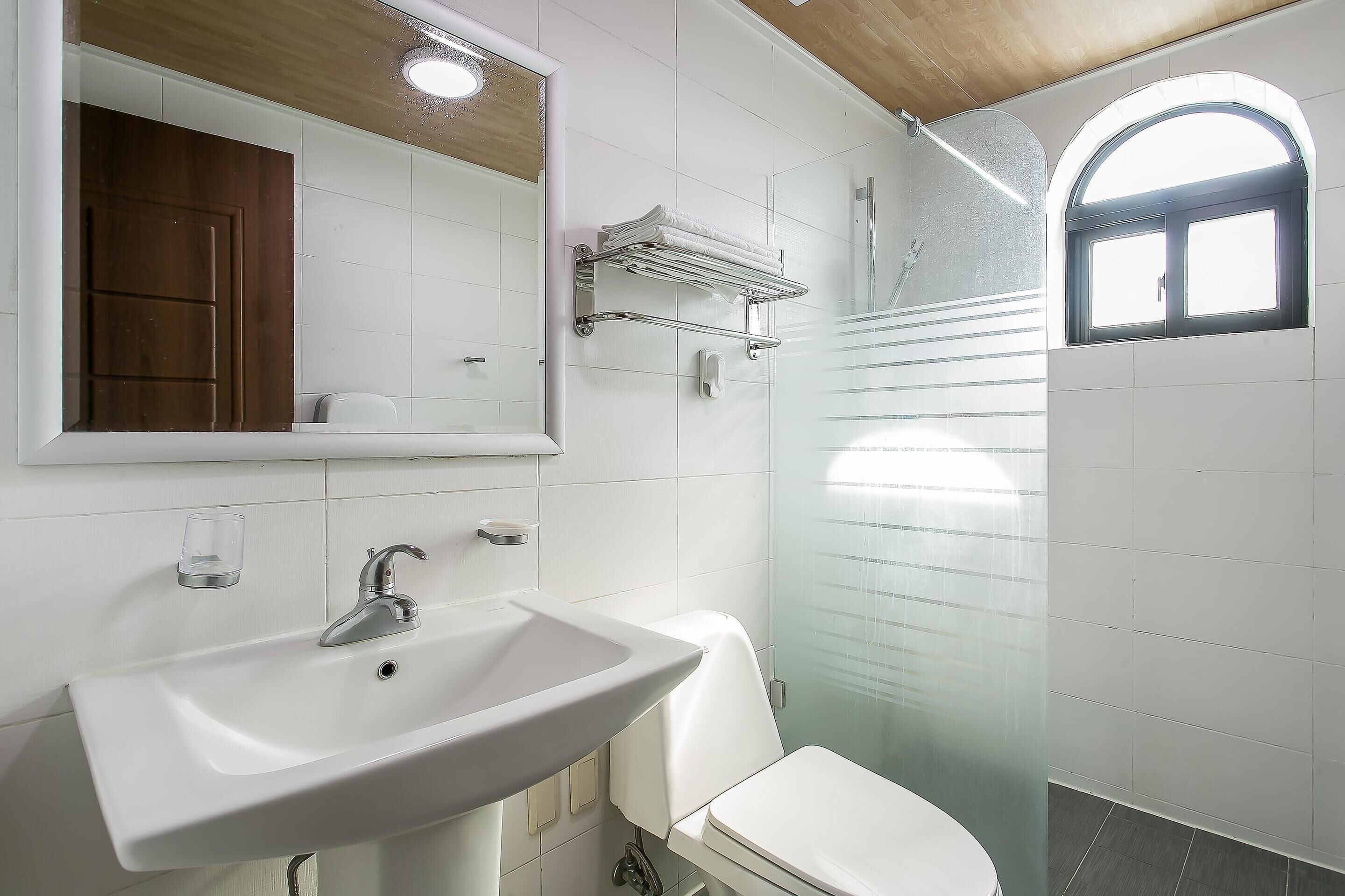 A modern bathroom with a white sink, mirror, and toilet. There is a glass shower enclosure and a small window.