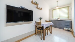 Une cuisine moderne avec un coin repas, un téléviseur fixé au mur, des étagères en bois et une fenêtre avec des stores.