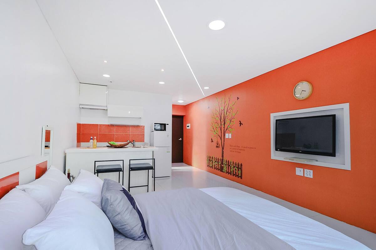 Ein modernes Hotelzimmer mit Bett, Fernseher, Küchenzeile und einer Wand mit einer Uhr und einem Baumgemälde.