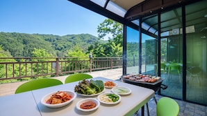 A dining area with a view of a forested hillside, a grill with food, and various dishes on the table. - Gapyeong Colline Pension (Gapyeong)