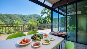 Un coin repas avec vue sur une colline boisée, un barbecue avec de la nourriture et différents plats disposés sur la table.