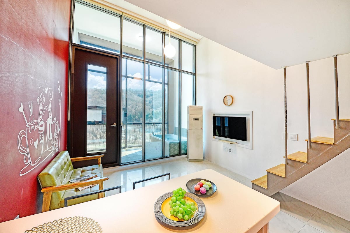 Ein modernes Interieur mit einem großen Fenster, einer Couch, einem Esstisch mit Obst und einer Treppe.