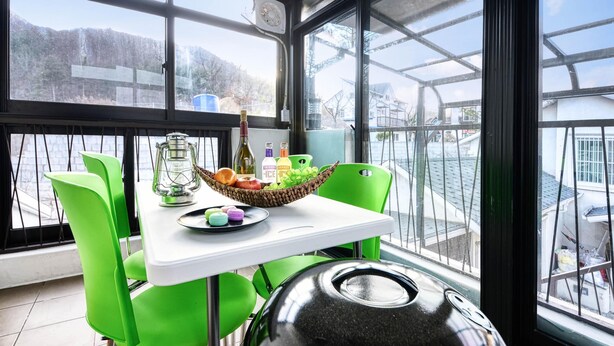 A room with a table set for dining, a green chair, a black grill, and a view of a snowy landscape through large windows. - Gapyeong Colline Pension (Gapyeong)