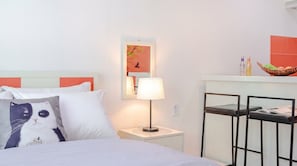 Une chambre d’hôtel avec un lit, une table de chevet avec une lampe, un miroir et une petite table sur laquelle se trouve un panier de fruits.