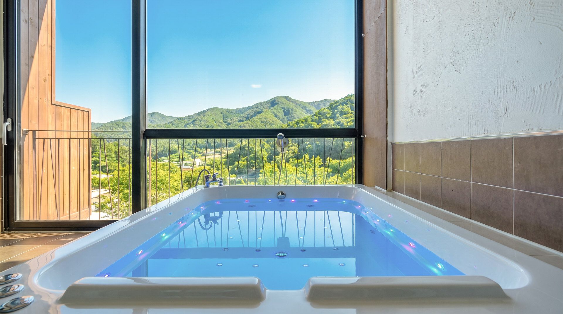 A spa room with a large bathtub, a view of green hills, and a balcony with a wooden railing.